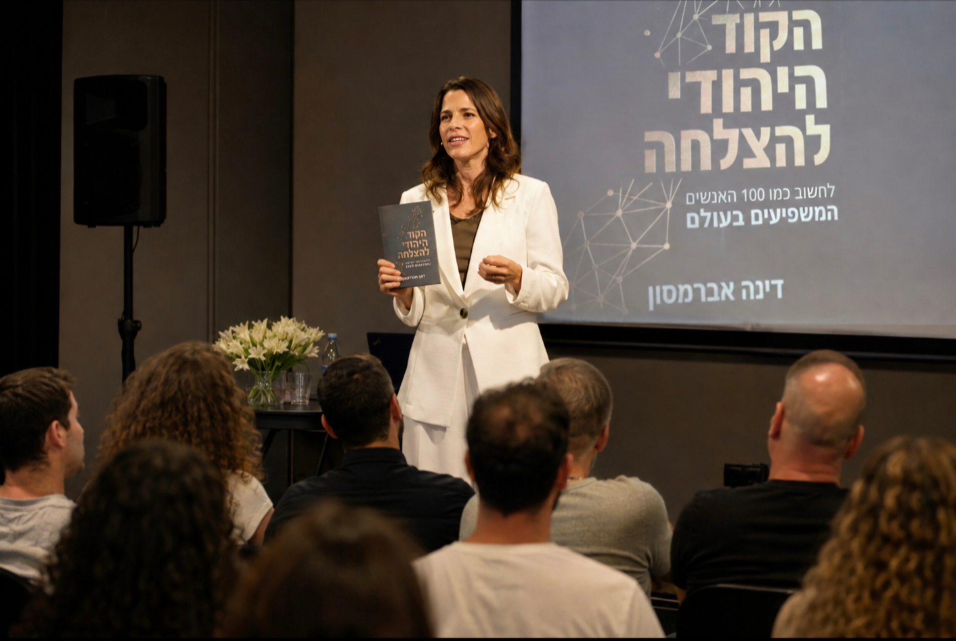 דינה אברמסון מרצה מול קהל ומחזיקה את הספר הקוד היהודי להצלחה